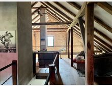 Holzgetäfelter Dachboden mit sichtbaren Holzbalken und einem Ziegelfußboden. Im Raum befinden sich eine Couch, ein kleines Fernrohr auf einem Stativ und eine gerahmte Schwarz-Weiß-Fotografie an der Wand. Ein Fenster wirft Tageslicht in die rustikal eingerichtete Umgebung.