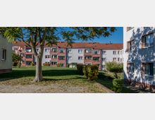 Mehrfamilienhäuser in einem Wohngebiet mit roten Dächern und hellen Fassaden, umgeben von Grünflächen, einzelnen Bäumen und Hecken. Ein gepflasterter Weg führt durch den Vordergrund.