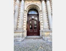 Großer historischer Eingang mit verzierten braunen Holztüren und einem halbkreisförmigen Oberlicht, flankiert von massiven steinernen Säulen. Die Fassade ist beige und mit architektonischen Verzierungen wie Reliefs und einer Hausnummer „4“ geschmückt, der Boden besteht aus Kopfsteinpflaster.