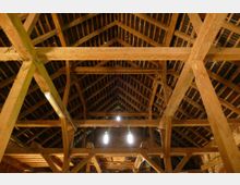 Holzstruktur eines Dachstuhls mit sichtbaren Balken und Sparren, die ein Giebeldach tragen. Tageslicht dringt durch Öffnungen im Dach hinein, und die Konstruktion zeigt eine traditionelle Zimmermannsarbeit.