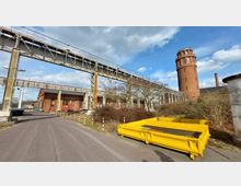 Industrielle Szenerie mit einem ziegelroten Wasserturm im Hintergrund, einem niedrigen Backsteingebäude, einer hohen metallischen Rohrbrücke und einer gelben Containerbox im Vordergrund. Die Umgebung ist von Sträuchern und einer asphaltierten Straße geprägt, während der Himmel klar bis leicht bewölkt ist.