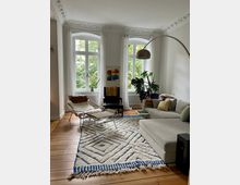 Wohnzimmer mit holzfarbenem Dielenboden, einem großen cremefarbenen Teppich mit blauen Mustern und einem grauen Ecksofa. Drei hohe Fenster mit weißen Vorhängen lassen viel Licht herein, ergänzt durch dekorative Stuckverzierungen an der Decke. Im Raum befinden sich eine Stehlampe, ein schwarzer Sessel, eine weiße Hängeliege und grüne Zimmerpflanzen.