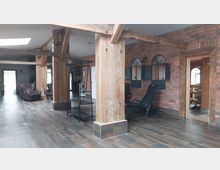 Großzügiger Innenraum mit sichtbaren Holzbalken, rotem Backsteinmauerwerk und modernen Fliesen. Der Raum enthält schwarze Stühle, ein metallisches Regal sowie eine geöffnete Tür, die in einen Bereich mit Saunaholzdekor führt.