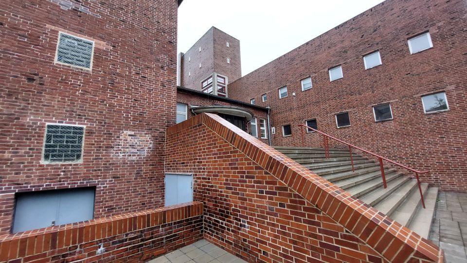 Backsteinarchitektur mit moderner Formsprache: Ein Gebäudekomplex aus rotem Klinkerstein mit Fenstern in unterschiedlichen Größen. Es sind mehrere Treppenaufgänge und eine erhöhte Eingangstür mit der Aufschrift „Stadtraum“ erkennbar, umgeben von klaren geometrischen Linien.