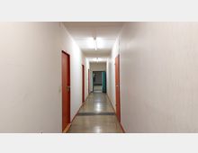 Langer, schmaler Flur mit weißen Wänden, rotem Türrahmen und mehreren geschlossenen Türen auf beiden Seiten; der Boden ist aus glänzendem Beton, und am Ende des Flurs ist eine geöffnete blaue Tür sichtbar.