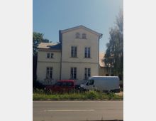 Zweistöckiges helles Wohngebäude mit Satteldach und mehreren rechteckigen Fenstern, an einer Straße gelegen. Im Vordergrund stehen ein roter Kleinwagen und ein weißer Lieferwagen, umgeben von vereinzeltem Grasbewuchs und einem Metallzaun.