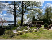 Außenbereich eines Cafés oder Restaurants mit Liegestühlen, Tischen und Stühlen in einem Garten. Große Bäume spenden Schatten, im Hintergrund liegt ein See mit Steg, und der Himmel ist bewölkt mit sonnigen Abschnitten.