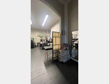 Ein helles Studio mit dunklem Fliesenboden und weißen Wänden, eingerichtet mit einem Massagetisch, Pflanzen sowie diversen Schränken und Kisten. Im Hintergrund hängen gerahmte Bilder an den Wänden, und leuchtende Deckenlampen erhellen den Raum.