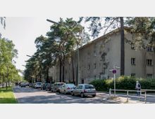 Straßenszene mit geparkten Autos entlang eines Gehwegs, der von hohen Kiefern und einem langgestreckten, vierstöckigen Wohngebäude mit schlichtem, beigem Putz gesäumt ist; im Vordergrund ein Straßenschild und eine Fußgängerin.