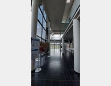 Moderner Innenbereich mit hohen Decken, großen Glasfenstern und weißen Säulen. Im Vordergrund stehen Anschlagtafeln, während der Blick durch eine verglaste Eingangstür ins Freie fällt.
