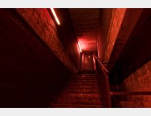 Eine enge Treppe in einem Betongebäude mit rotem, gedämpftem Neonlicht, das die Wände und Stufen beleuchtet. Am oberen Ende der Treppe befindet sich eine Metalltür mit Graffiti.