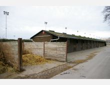 Holzstallgebäude mit langen Reihen von Stalltüren unter einem flachen Dach, das teilweise mit Gras bewachsen ist. Im Vordergrund befinden sich Holzzäune und ein Bereich mit Stroh, im Hintergrund sind Flutlichtmasten und ein städtisches Umfeld sichtbar.