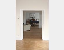 Blick durch eine geöffnete Doppeltür in ein Esszimmer mit Holzboden. Im Hintergrund steht ein gedeckter Esstisch mit blauer Tischdecke, umgeben von Stühlen, und darüber hängt ein gerahmtes Bild an der Wand.