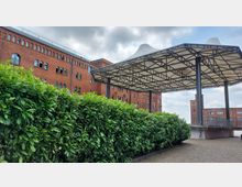 Ziegelgebäude mit mehreren Stockwerken und runden Fenstern im Hintergrund, vorne ein modernes, großes Dach mit Metallkonstruktion und weißen Spannstoffen, umgeben von einer dichten Hecke und Pflastersteinen.