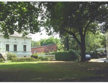 Ein parkartiges Gelände mit Rasenfläche und einem großen Baum im Vordergrund. Links ein weißes Gebäude im klassizistischen Stil mit verzierten Fenstern, rechts eine Backsteinmauer, ein Torbogen mit Schriftzug und ein geparktes Auto.