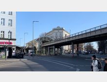 Straßenkreuzung in einer Stadt mit erhöht verlaufender U-Bahn-Brücke und Bahnhofseingang im Zentrum, flankiert von einem historischen Gebäude mit heller Fassade und einem Eckgebäude mit dem Schriftzug „QUE PASA“. Im Hintergrund Bäume und eine gerade, mit Autos befahrene Straße.