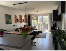 Offener Wohn- und Essbereich mit Kücheninsel im Vordergrund, Esstisch und Sofa; über dem Tisch hängen drei geflochtene Pendelleuchten. Große bodentiefe Glastüren führen auf einen Balkon, rechts eine dunkle Einbauwand mit Stauraum, dazu Pflanzen und ein Teppich auf hellem Boden.