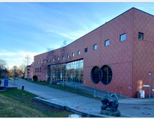 Backsteinbau einer modernen Einrichtung mit rechteckigen und kreisförmigen Fensterformen, großer Glasfassade und einer Skulptur im Vordergrund. Das Gebäude ist von einem gepflasterten Platz sowie einem Grünstreifen mit Bäumen umgeben.