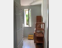 Helles Badezimmer mit einem geöffneten Fenster, durch das grünes Blattwerk sichtbar ist. Links ein weißer Handtuchheizkörper und rechts ein Holzregal neben einem Waschbecken mit metallener Armatur; ein gelber Tritthocker steht vor dem Fenster.