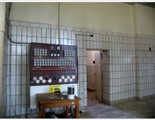 Ein technischer Raum mit vollständig gefliesten Wänden in hellen Farben. Links ist ein alter Schaltschrank mit zahlreichen Schaltern, Knöpfen und Anzeigen zu sehen, während rechts eine geöffnete Tür zu einem angrenzenden Raum führt. Ein kleiner Holztisch mit verschiedenen Gegenständen steht davor.