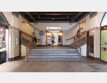 Eingangsbereich einer U-Bahn-Station mit hell gefliesten Wänden, einer mittigen Treppe, die zu zwei Aufgängen mit roten Ticketautomaten führt. Links und rechts befinden sich Geschäfte und zwei symmetrische Treppen mit Holzgeländern.