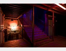 Metalltreppe in einem industrialen Kellergang mit sichtbarer Stahlkonstruktion, beleuchtet von rotem und violettem Licht. Graffiti und Schilder mit Wegweisern sind an den rauen Betonwänden angebracht.