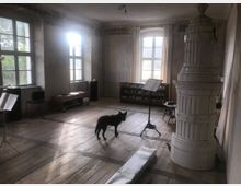 Ein heller Raum mit großen Fenstern, einem Holzboden und einem weißen Kachelofen. Im Raum stehen Notenständer, ein Regal mit Geschirr sowie weitere Möbel; ein schwarzer Hund befindet sich in der Mitte.