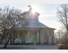 Runder Pavillon mit grün-goldener Fassade und reichem Dekor im Rokoko-Stil, umgeben von kahlen Bäumen bei Sonnenlicht. Oben auf dem Dach ist eine vergoldete Figur mit Sonnenschirm sichtbar.