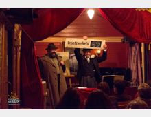 Innenansicht einer kleinen, rustikalen Bühne mit roten Vorhängen und warmem Licht. Zwei Männer in historischer Kleidung, einer mit einem Mantel und Hut, der andere mit Anzug und Blume im Knopfloch, präsentieren ein Schild mit der Aufschrift „Ersatzverkehr 69“ vor einem sitzenden Publikum.