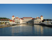 Großes Hotelgebäude im modernen Baustil mit gelben und weißen Fassaden sowie roten Dächern, das an einem Wasserkanal liegt. Mehrere weiße, geschwungene Brücken führen über das Wasser, während im Hintergrund ein Wald zu sehen ist.