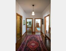 Heller Flur in einer Wohnung mit Holzdielenboden, orientalischem Läuferteppich und zentraler Pendelleuchte. Mehrere Türöffnungen mit Holzrahmen führen in angrenzende Räume; rechts steht ein Holztisch mit Stuhl, links eine Glastür.