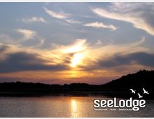 Sonnenuntergang über einem See mit reflektierendem Licht auf der Wasseroberfläche, im Hintergrund eine bewaldete Uferlinie und teilweise bewölkter Himmel. In der unteren rechten Ecke befindet sich ein Logo mit der Aufschrift „seeLodge“.