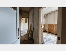 Ein verlassener, heruntergekommener Innenraum mit abblätternder Tapete an den Wänden und beschädigtem Bodenbelag. Im Vordergrund ein Flur mit Blick in einen Raum mit kariertem Fliesenboden und teils abgefallenen Wandpaneelen.