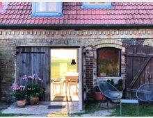 Backsteinfassade eines Hauses mit roten Dachziegeln, einer Holztür, einem Fenster und sichtbaren Holzakzenten. Im Vordergrund stehen Blumentöpfe mit rosa Blumen, daneben befinden sich zwei Metallstühle und ein kleiner Tisch auf einer Grasfläche.