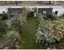 A backyard garden with a rectangular lawn bordered by bushes and small trees. In the background, there is a modern, single-story building with a flat roof, dark gray and white walls, and large windows. A soccer goal and basketball hoop are visible in the yard, with a park and trees beyond the property.