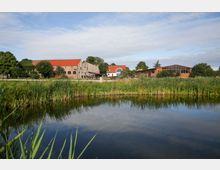 Gebäudeensemble mit roten Ziegeldächern und moderner Glasfassade, umgeben von Grünflächen und Bäumen, im Vordergrund ein Schilf gesäumter Teich unter blauem Himmel.