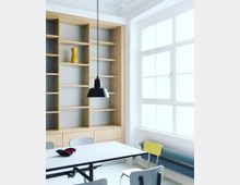 Ein heller Raum mit einer großen Fensterfront, einem weißen Tisch und grauen sowie gelben Stühlen. Im Hintergrund steht ein Regal aus hellem Holz mit offenen Fächern und Schubladen, darüber hängt eine schwarze Hängelampe.