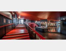 Moderner Innenraum eines Gebäudes mit roten Treppen und einer Holzdecke. Links führt eine Rampe mit Geländer, rechts befindet sich ein Tresen, und dahinter sind Wände mit beleuchteten Bildern sowie ein glatter, dunkler Boden sichtbar.