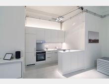 Moderne, minimalistische Küche mit weißen Schränken, integriertem Backofen, Kochfeld und Dunstabzugshaube. Eine freistehende Kücheninsel sowie dezente Wanddekoration und sichtbare Kabelverrohrungen ergänzen das helle Raumdesign.