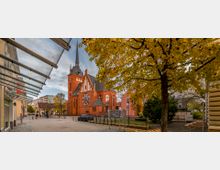 Rote Backsteinkirche mit spitzem Turm und großen, runden Fenstern, umgeben von Bäumen mit herbstlich gelbem Laub; im Vordergrund Gehwege und einige Autos.