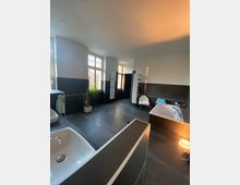 Moderner, geräumiger Badezimmerbereich mit dunklen Fliesen, weißen Waschbecken, einer Eckbadewanne und hohen Fenstern, die Tageslicht hereinlassen; eine Topfpflanze und ein Handtuchstuhl ergänzen die Einrichtung.