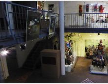 Innenansicht eines Museums mit einem Schwerpunkt auf Puppen oder Marionetten. Zu sehen sind zwei Etagen mit einer metallischen Galeriebrüstung, Puppen in gläsernen Vitrinen sowie Wandposter und dekorative Ausstellungsobjekte.