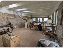 Großer, offener Raum mit Sichtmauerwerk an den Wänden, einer Holzdecke mit weißen Balken und Fenstern auf der rechten Seite. Der Raum ist spärlich eingerichtet mit Holzmöbeln, einem Holzofen mit Edelstahlrohr und diversen Gegenständen wie Kisten, Sofas und Küchenutensilien.