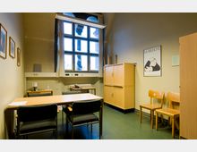 Kleines Büro oder Besprechungszimmer mit einem großen Fenster, einem Holztisch und mehreren Stühlen. An den Wänden hängen Bilder, ein Poster mit der Aufschrift "Loriot", und es gibt Holzschränke sowie kleinere Tische mit einer Kaffeemaschine und einem Telefon.