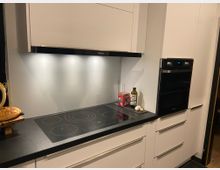 Moderne Küche mit weißen Einbauschränken, schwarzer Arbeitsplatte und einem Induktionskochfeld. Rechts befinden sich ein Einbaubackofen und eine beleuchtete Dunstabzugshaube über dem Kochfeld; auf der Arbeitsplatte stehen Ölflasche und Gewürze.