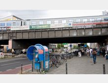 Straßenszene mit einer erhöhten Eisenbahnbrücke, auf der ein Zug fährt, darunter Gehweg und Fahrbahn. Im Vordergrund steht eine blaue öffentliche Telefonzelle, daneben eine Reihe abgestellter Fahrräder; mehrere Passanten bewegen sich entlang des Gehwegs.