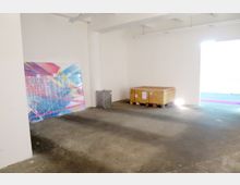 Ein leerer Raum mit weiß gestrichenen Wänden und Betonboden. Im Vordergrund steht eine Holzkiste auf einer Palette, daneben befindet sich ein verpacktes Kunstwerk mit bunten, geometrischen Mustern an der Wand.