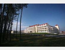 Großes, mehrstöckiges Hotelgebäude mit rotem Satteldach und zahlreichen Balkonen, umgeben von einer grünen Wiese und hohen Bäumen im Vordergrund. Der Himmel ist klar und blau, und vor dem Gebäude sind Flaggen aufgestellt.