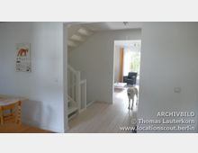 Heller Wohnraum mit offener Treppe, weißen Wänden und hellem Holzfußboden. Im hinteren Bereich befindet sich ein Wohnzimmer mit großen Fenstern, gelben Vorhängen, einem schwarzen Sessel und einem weißen Tisch. Ein Hund steht im Flur.