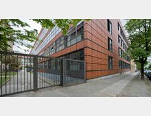 Moderne Bürogebäude mit Fassade aus roten Kacheln und großen Glasfenstern. Vor dem Gebäude befindet sich ein gepflasterter Bürgersteig, ein Metalltor und angrenzende Grünflächen mit Bäumen.